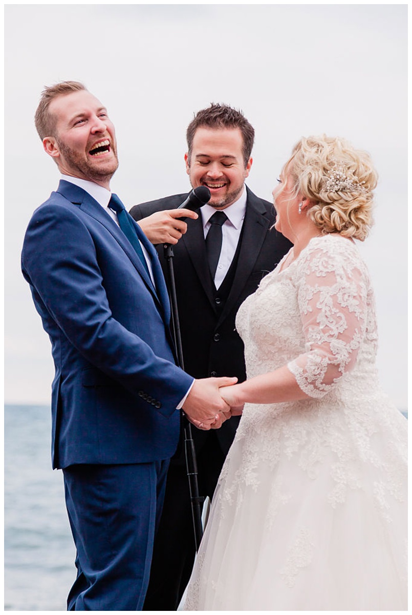 redheaded groom in a blue suit laughs uproariously at something the officiant says during the wedding ceremony on the beach