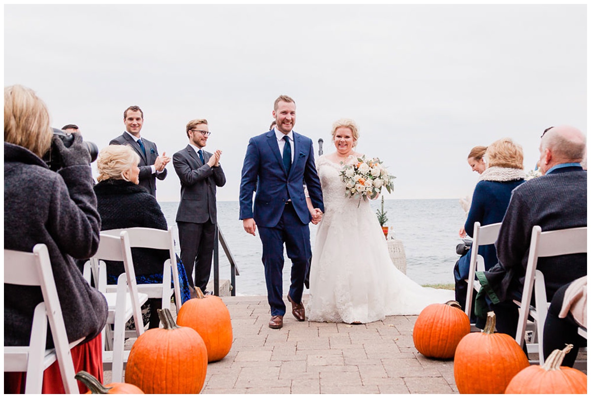 a redheaded groom in a blue suit and his happy blonde bride walk down the aisle lined with pumpkins as husband and wife 