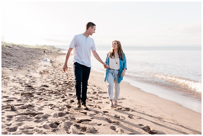 a couple walks along the beach, he is wearing a white t-shirt and jeans, she is wearing jeans, a white shirt and a turquoise sweater. They are smiling and laughing showing off their engagement outfits. Photos by Comfort and Cashmere