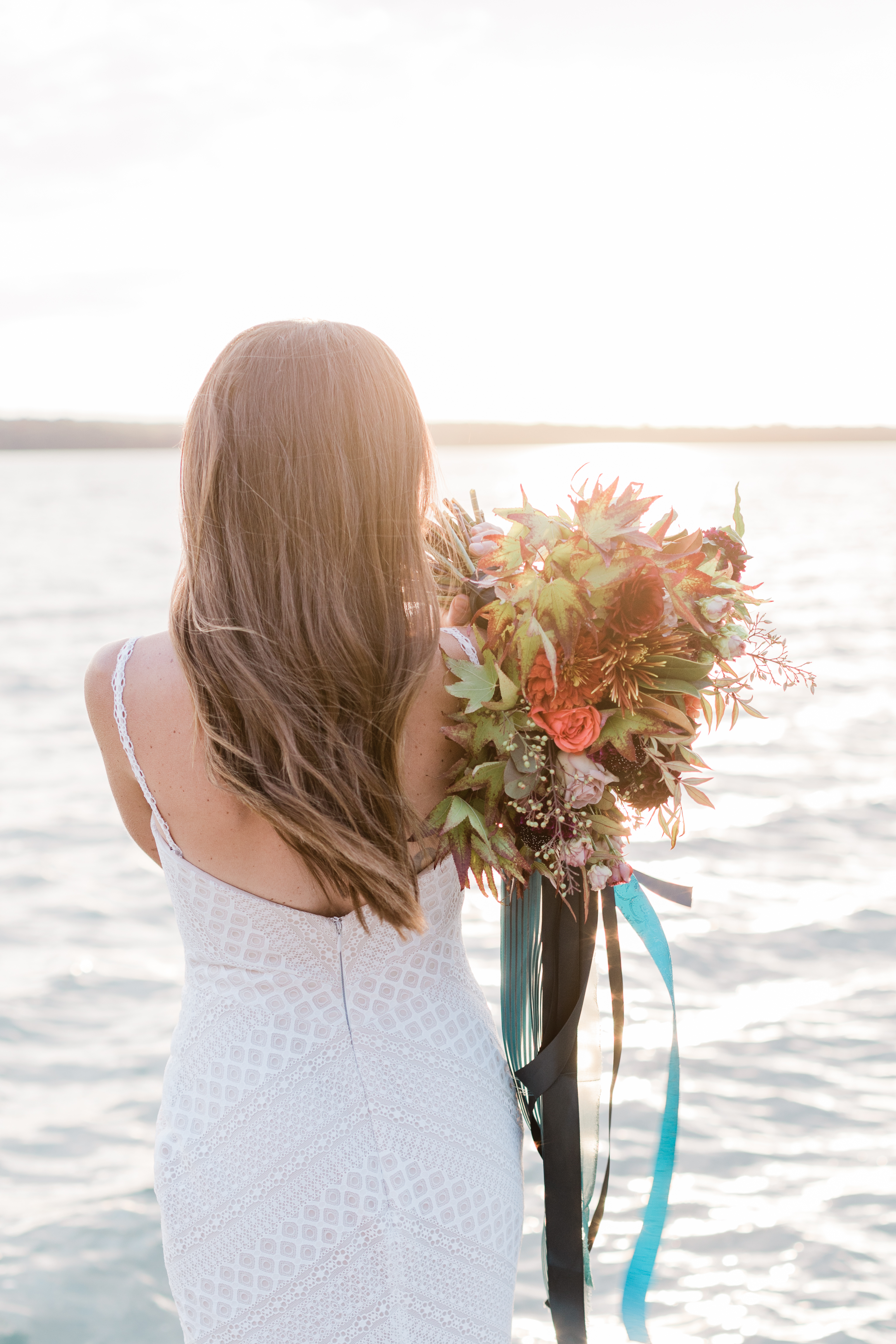 brunette bride holds huge bouquet of seasonal flowers over her shoulder and looks out at ocean.