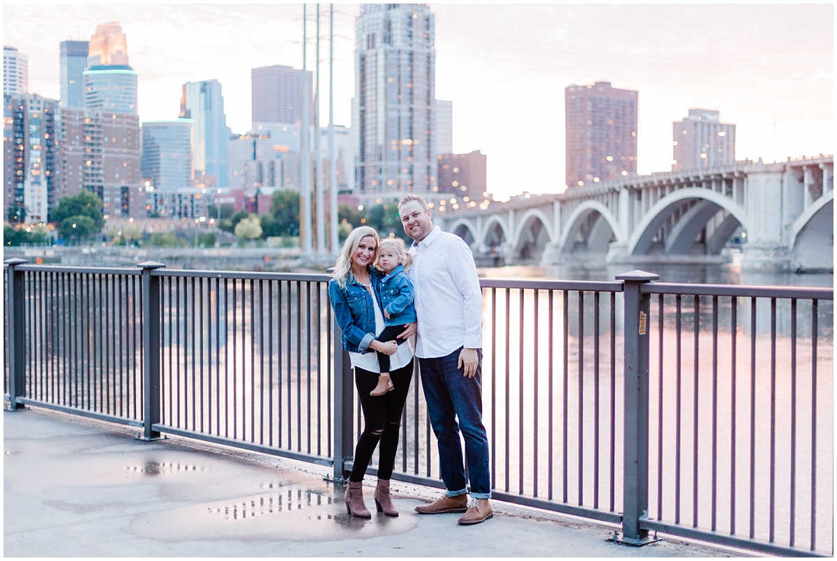 Family photo of man, woman and their blonde toddler in front of the Mississippi River in St. Anthony Main in Minneapolis