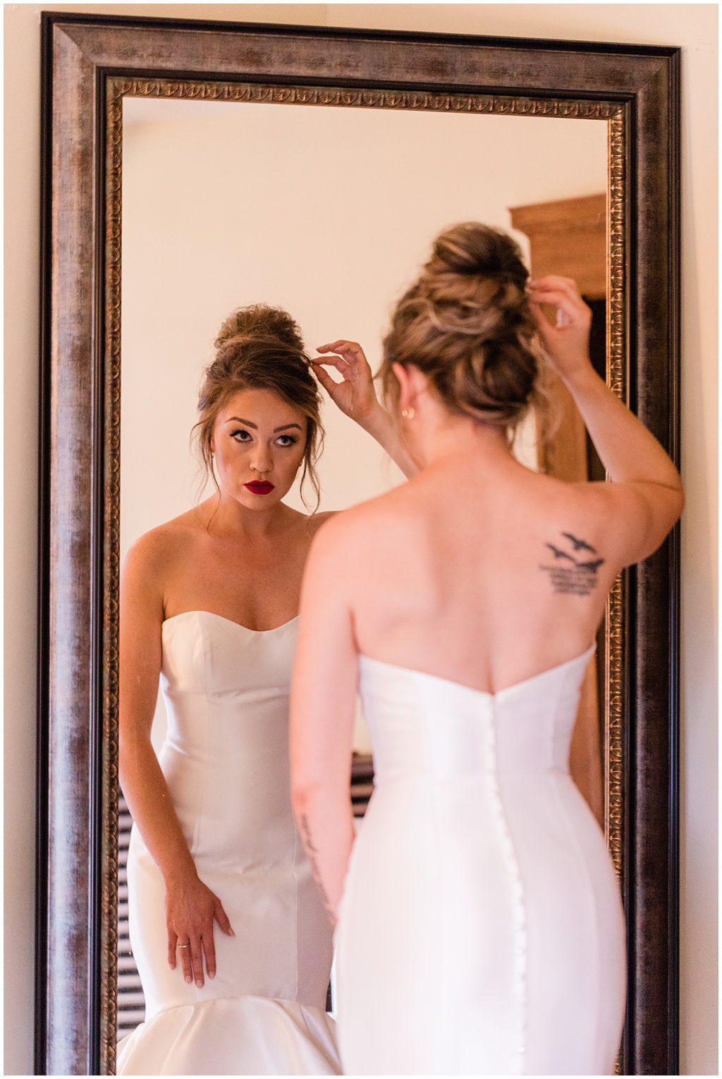 bride with a shoulder tattoo of birds and words wearing a strapless gown checks her hair in a full length mirror which also shows off the mermaid style of her dress and flattering figure