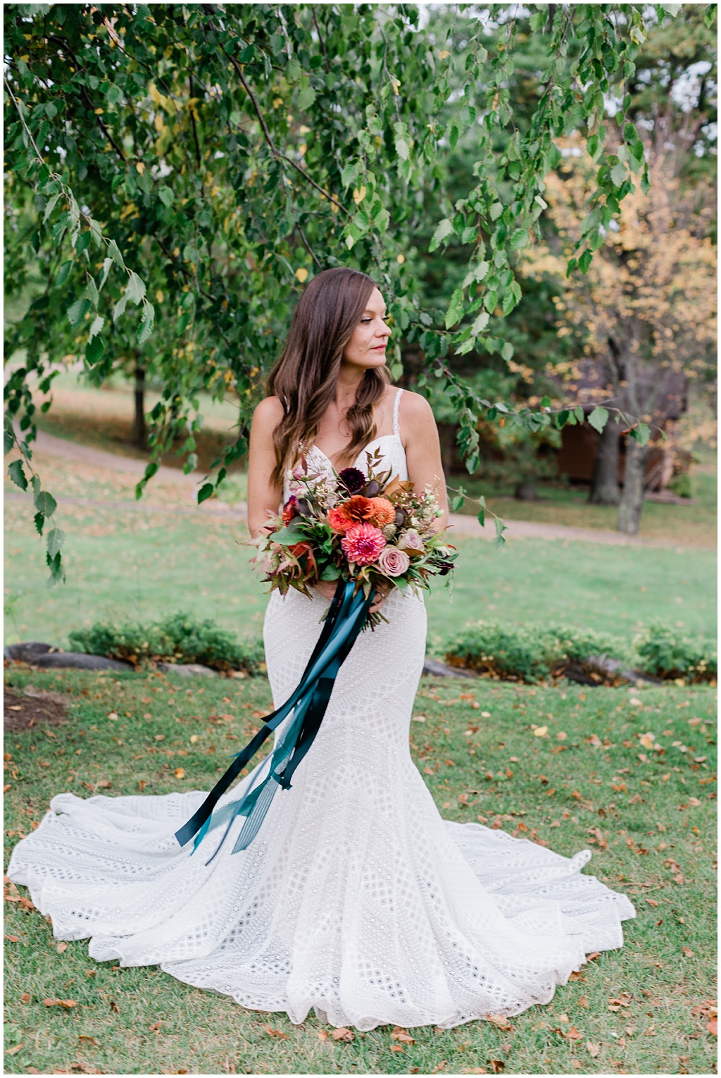 full length portrait of a long-haired, brunette bride holding a bouquet of seasonal summer flowers under a canopy of green leaves