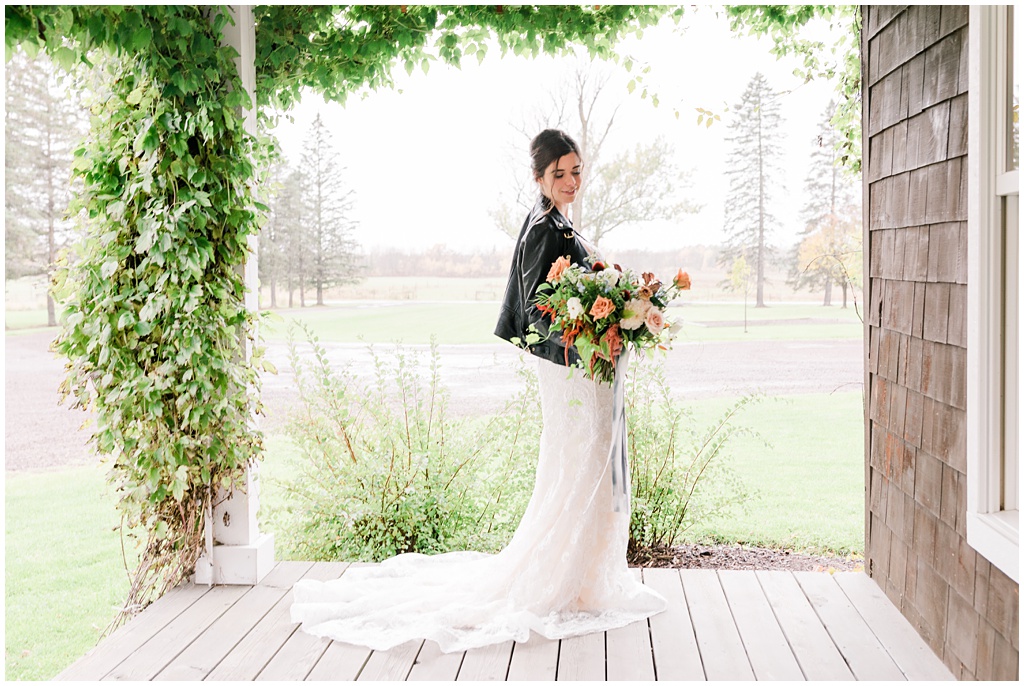 young bride wearing a leather jacket holds a giant bouquet of summer seasonal flowers while standing on a porch framed by green vines with a lake in the background