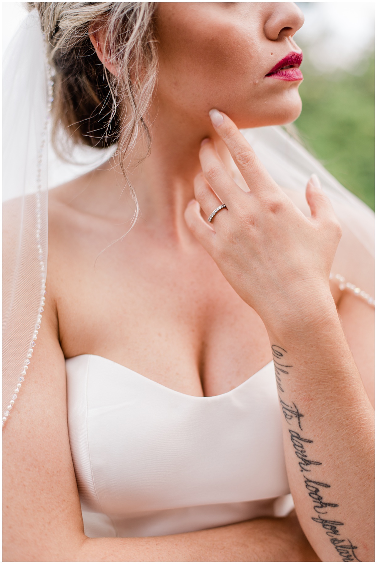 bride wearing a simple gown with an up do, runs a slender finger along the side of her face and displays the writing of her tattoos on her arm and a pop of purple lipstic. Dear Bride series on wedding planning