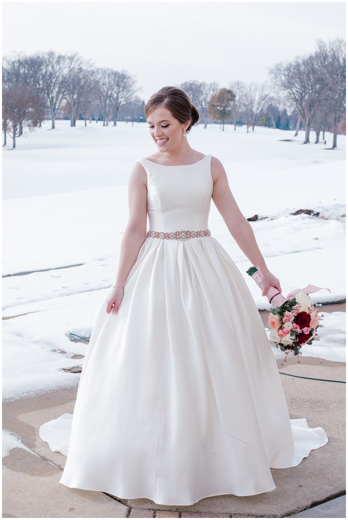Bride wearing a simple, yet elegant princess dress with a pink belt made of crystals, holds her bouquet and looks down with a winter wonderland behind her.