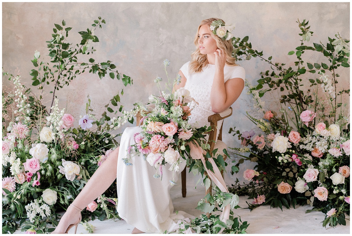 gorgeous blonde bride sits among a huge amount of gorgeous wedding flowers filled with colorful roses and greenery