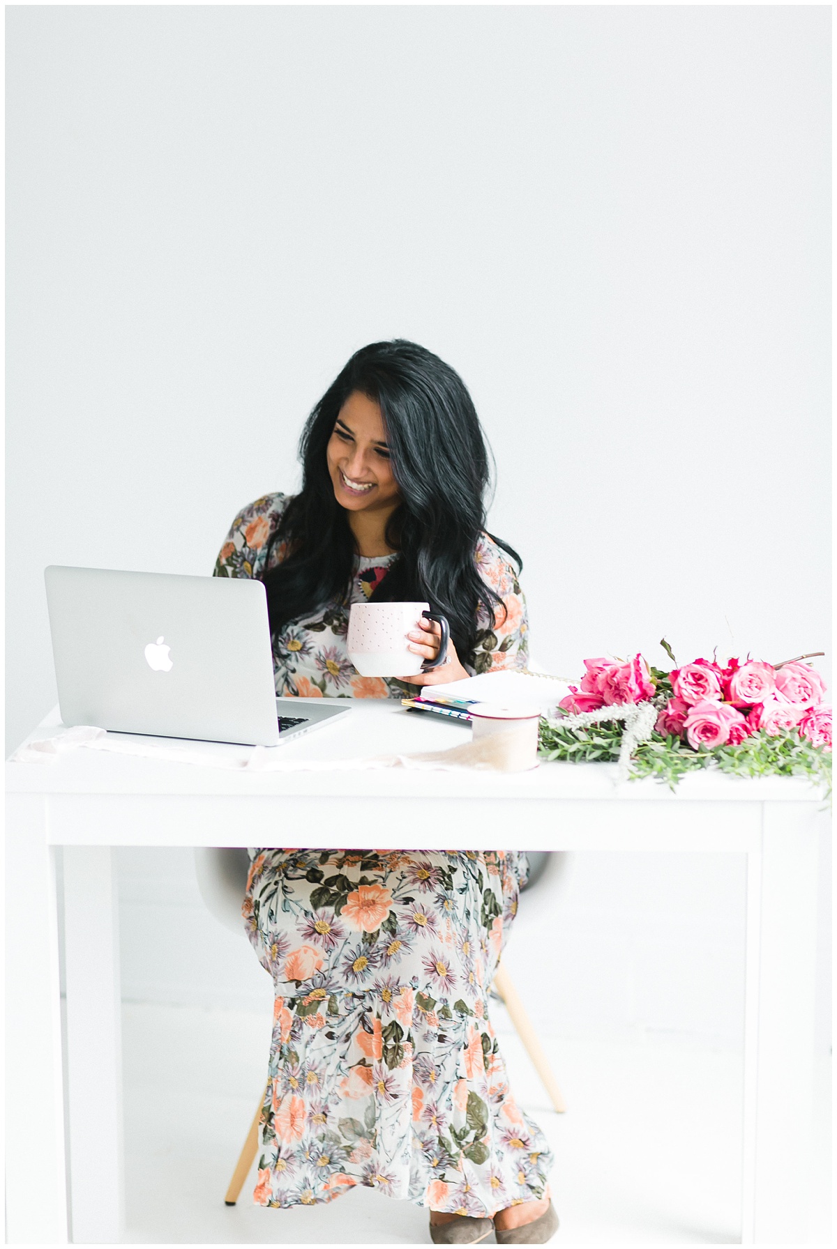 smiling photographer wearing a gorgeous floral print dress sits at a desk with coffee and her laptop, smiling and planning social posts with Planoly