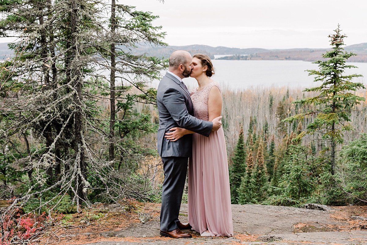A man in a gray suit kisses his bride wearing a long, pink dress as they stand on a mountain overlooking a lake for their elopement photos.