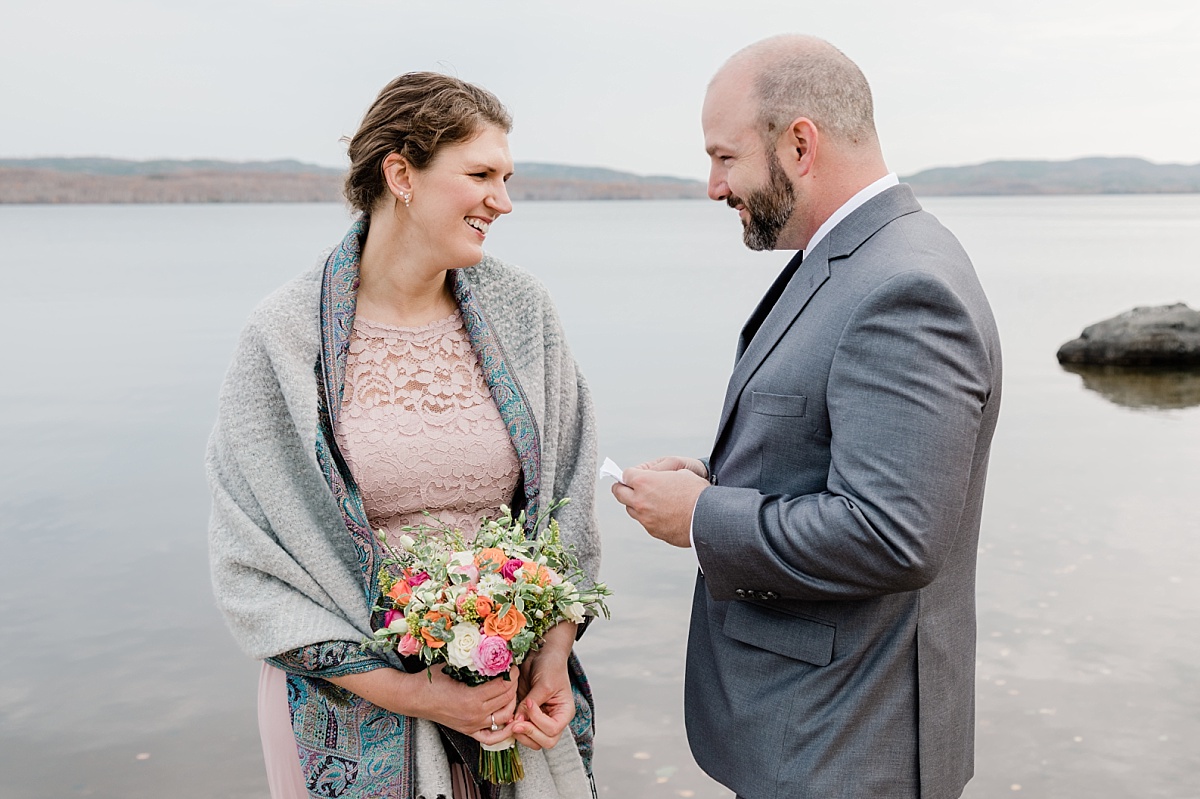A couple smiles at each other while reading vows in front of a lake during their elopement with Comfort and Cashmere Images.