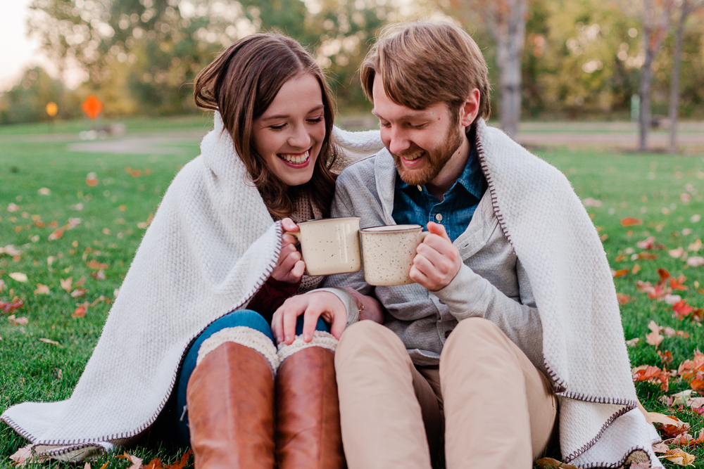 A smiling Caucasian couple sitting next to each other on the grass in the fall, wrapped in a blanket and clinking mugs during an engagement shoot with Comfort and Cashmere Images.