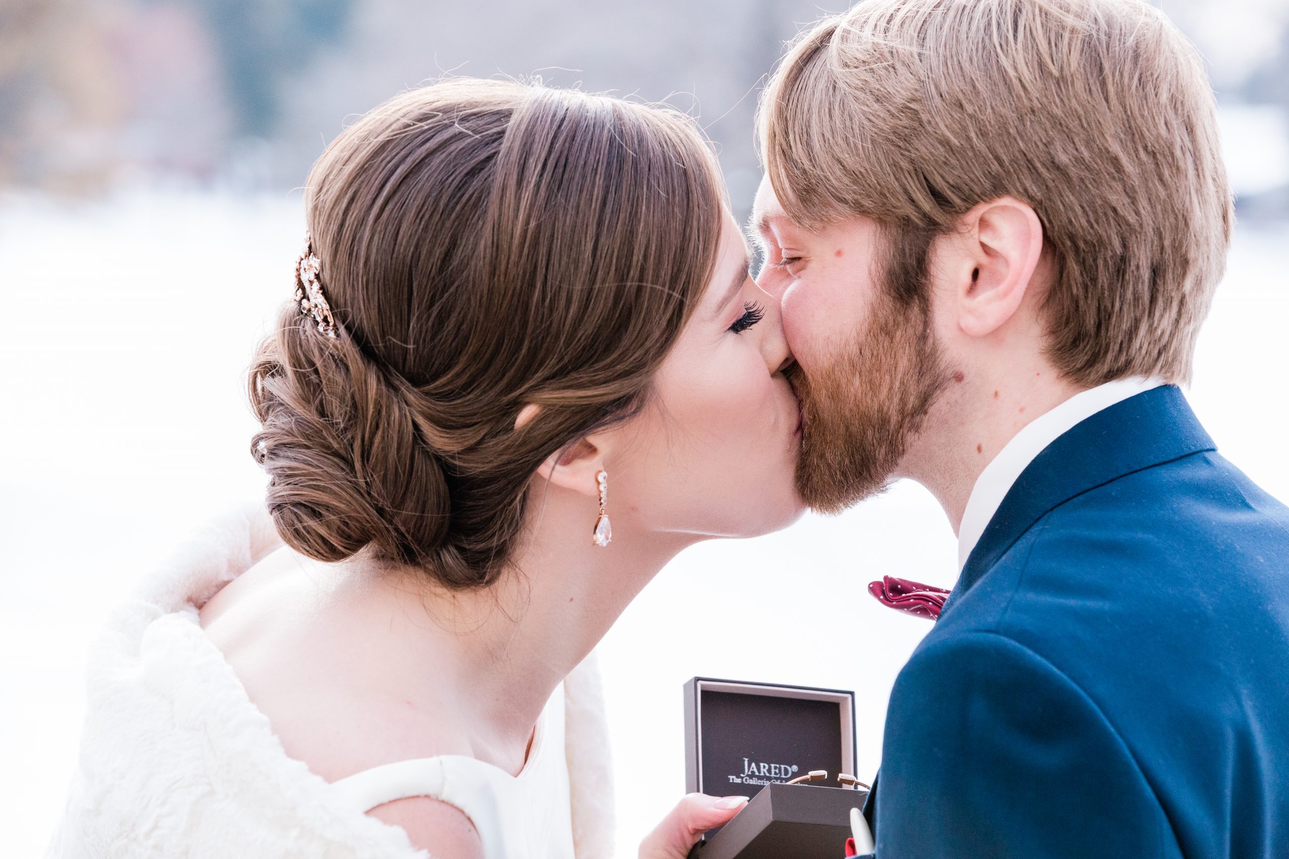 A brunette bride in a white dress and teardrop earrings kisses her bearded groom in a blue tux for winter outdoor wedding photos with Comfort and Cashmere Images in Minnesota.