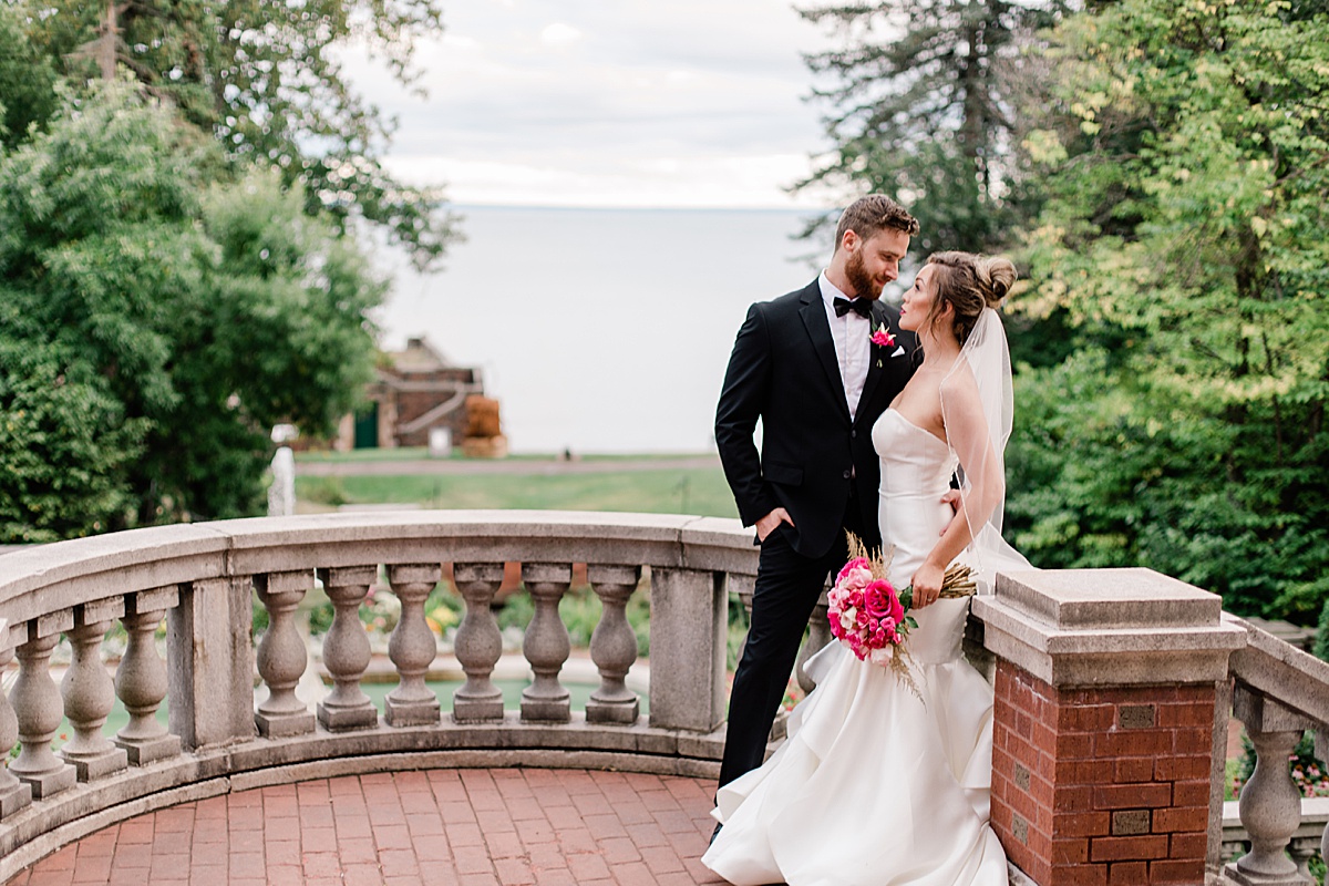 bride and groom stand on a red brick terrace leaning against an ornate cement bannister with a gorgeous lake behind them at Glensheen Mansion during their intimate wedding. 