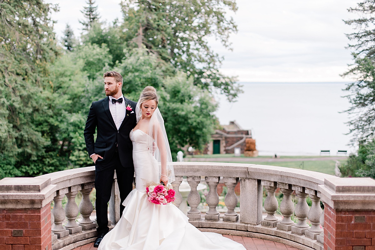 A bride leans against her groom on a castle terrace looking down at her pink bouquet in this styled luxury wedding shoot with Rohana Olson