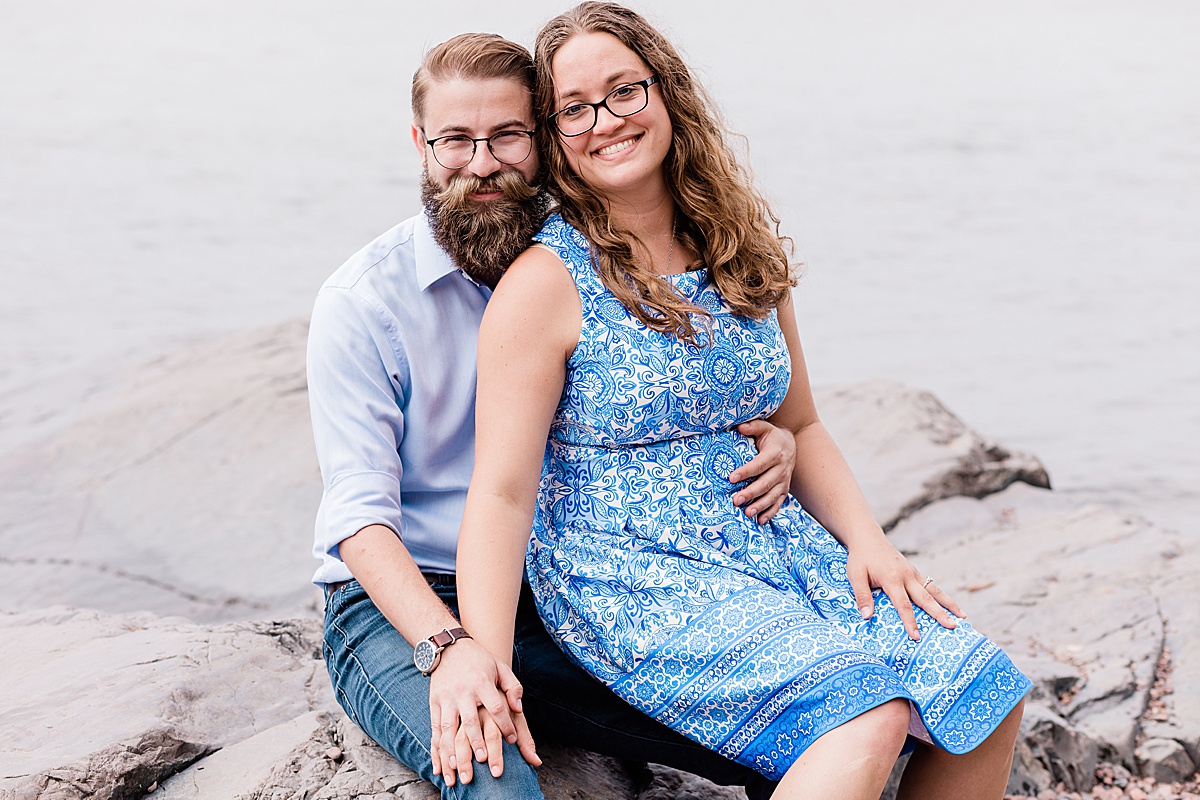woman wearing a pretty blue and white print dress sits on smiling fiance's lap riverside in Duluth, Minnesota during an engagement session with Comfort & Cashmere images