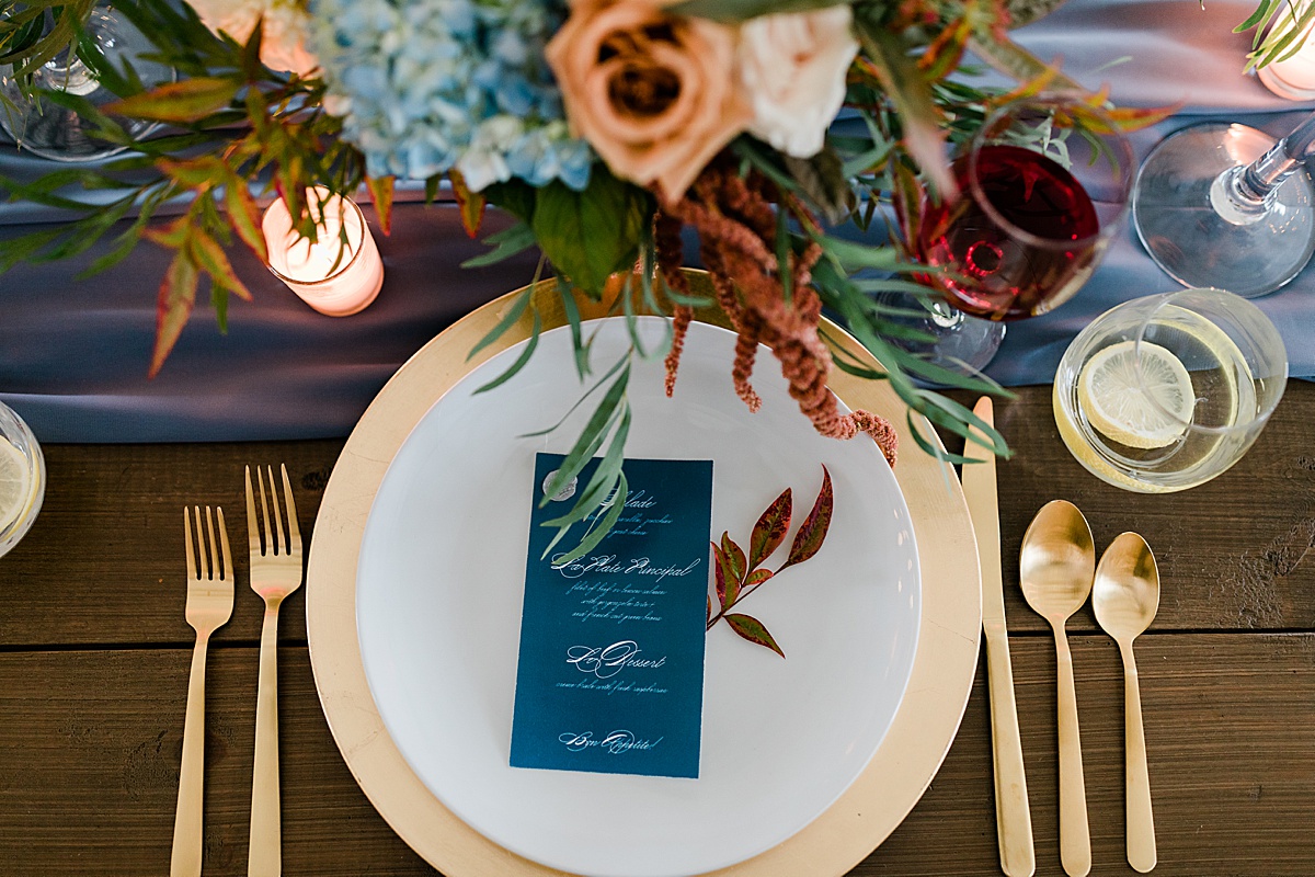 a gold-rimmed plate and gold utensils framed by blue and orange flowers and a blue menu with white writing and fall foliage demonstrate how to add luxury to your wedding