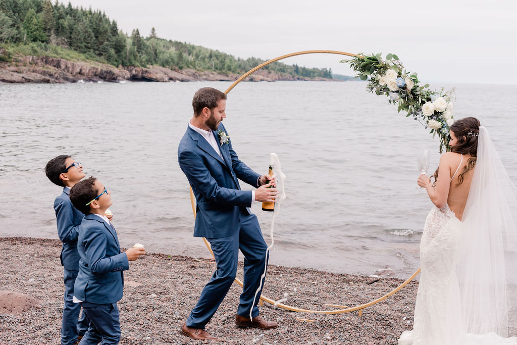 groom pops a bottle of champagne at the beach while he and his new bride celebrate all their reasons for eloping at Sugar Loaf Cove on Lake Superior