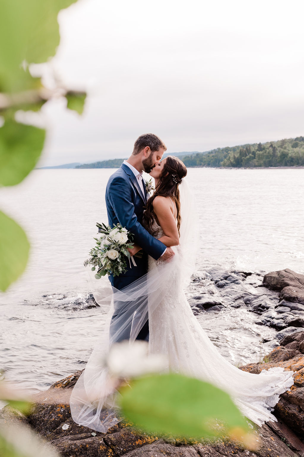foliage frames a newlywed couple as they kiss at the edge of Lake Superior at Sugar Loaf Cove in Minnesota with Comfort & Cashmere Images