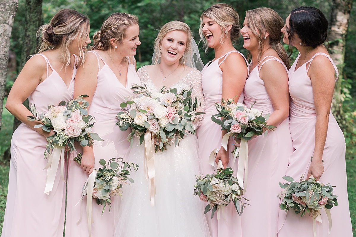 A bridal party in blush pink dresses surrounds the beautiful bride while holding bouquets and laughing together during a lovely Minnesota wedding.