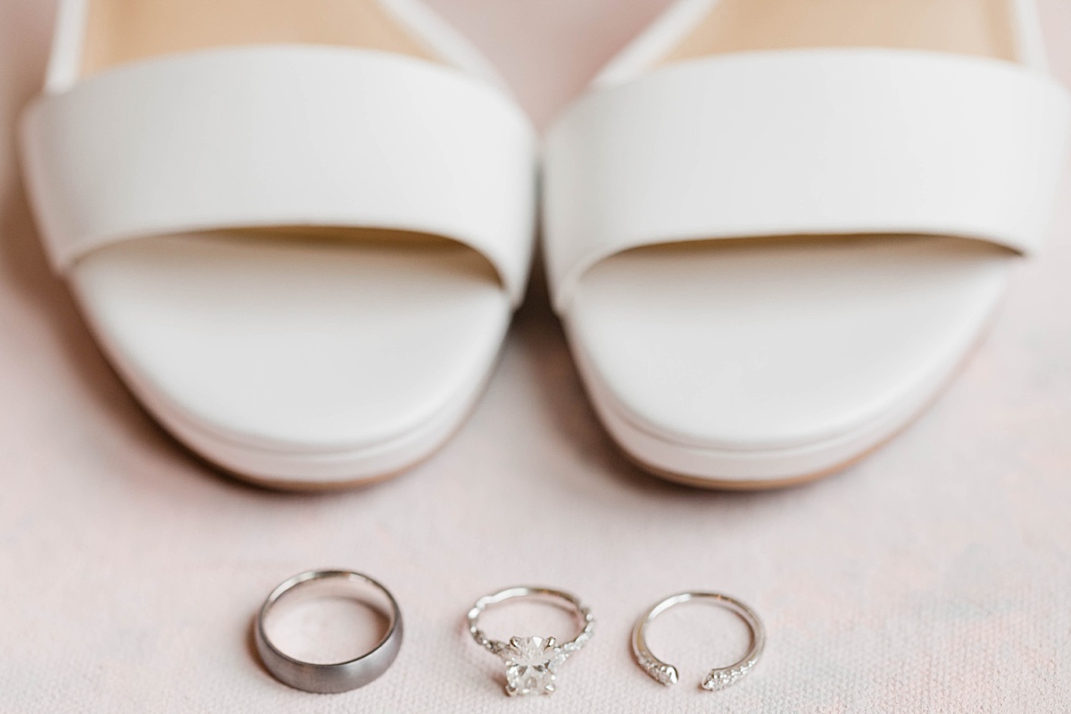 The toe area of Michael kors wedding heels with a man's wedding band, diamond engagement ring and diamond wedding band laid out in front of them